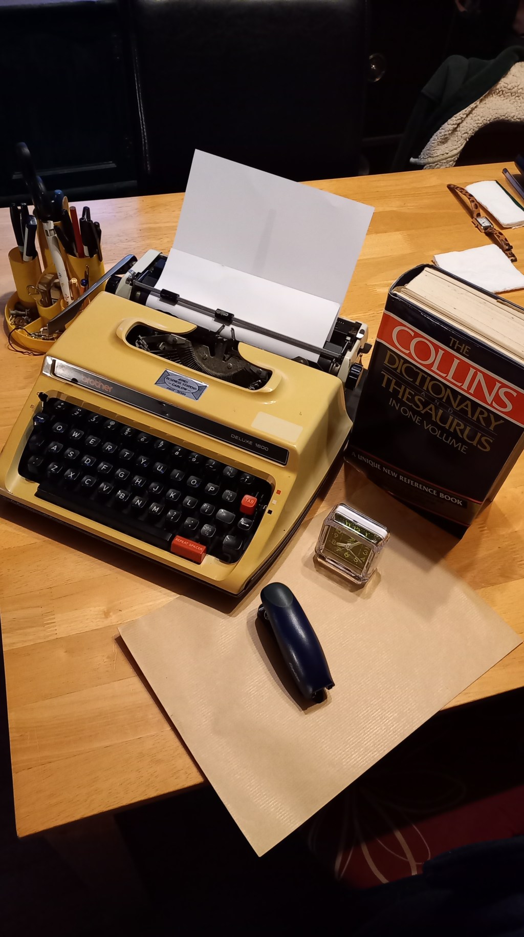 colour photograph showing a manual typewriter with a blank piece of paper inserted in the top; a hard-copy dictionary, analogue clock, stapler, brown envelope, and desk tidy containing pens, eraser fluid, etc.